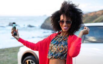 A smiling woman wearing sunglasses and a colorful crop top takes a selfie by the seaside, standing next to a white car with cliffs and ocean in the background.
