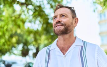A middle-aged man with a beard and short hair, wearing a white shirt with stripes, stands outdoors and looks thoughtfully into the distance. Blurred green trees and sunlight are in the background.
