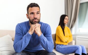 A man in a blue sweater sits on a couch, looking thoughtful with his hands clasped under his chin. Behind him, a woman in a yellow sweater sits with her arms crossed, facing away. Both appear upset.