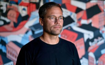 A man with short brown hair and round glasses wearing a black t-shirt, posed in front of a colorful abstract mural.