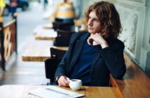 A person with wavy, shoulder-length hair sits at an outdoor café table, holding a cup of coffee and gazing thoughtfully to the side. A newspaper is on the table in front of them.