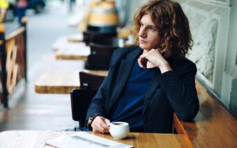 A person with wavy, shoulder-length hair sits at an outdoor café table, holding a cup of coffee and gazing thoughtfully to the side. A newspaper is on the table in front of them.