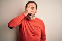 A man wearing a red sweater looks surprised, holding one hand over his face with fingers spread to reveal one eye, and his mouth open in shock against a plain light background.