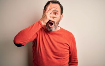 A man wearing a red sweater looks surprised, holding one hand over his face with fingers spread to reveal one eye, and his mouth open in shock against a plain light background.
