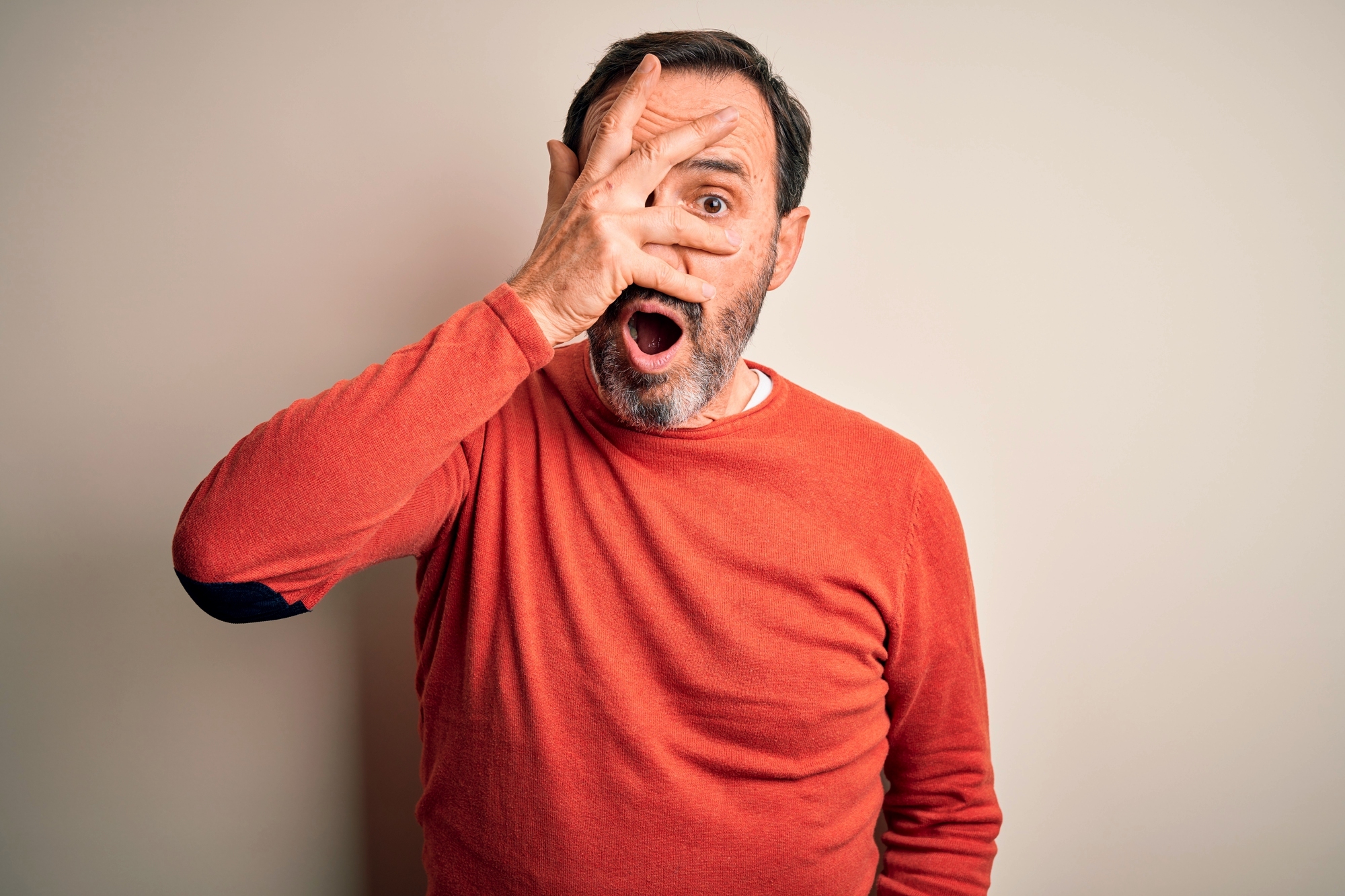 A man wearing a red sweater looks surprised, holding one hand over his face with fingers spread to reveal one eye, and his mouth open in shock against a plain light background.