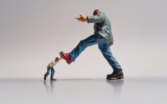 A small figurine in a white shirt and blue jeans pushes against the leg of a much larger figurine in a green jacket and boots, creating a dramatic size contrast on a plain background.