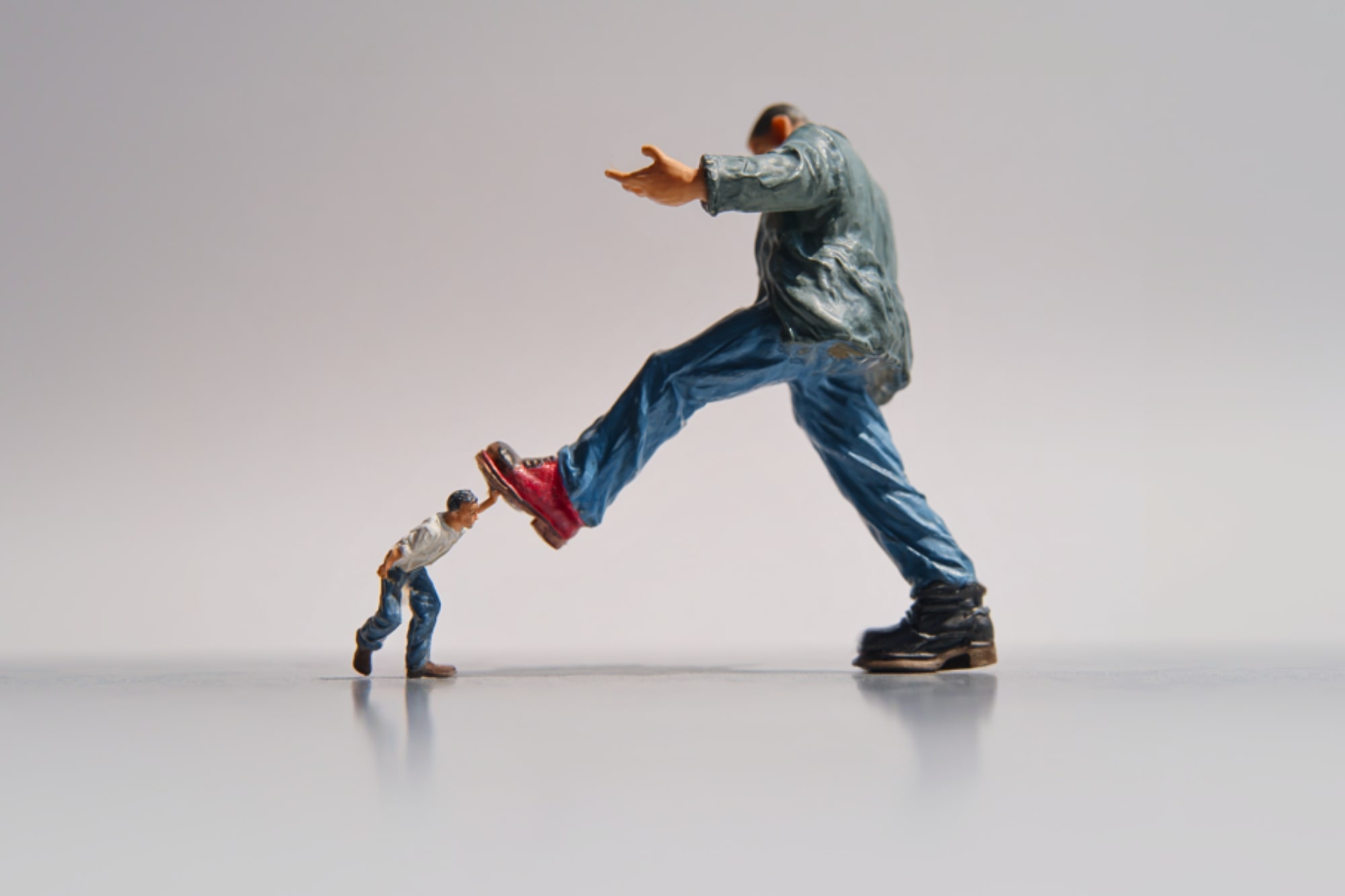 A small figurine in a white shirt and blue jeans pushes against the leg of a much larger figurine in a green jacket and boots, creating a dramatic size contrast on a plain background.