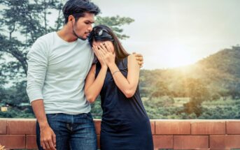 A man in a white shirt comforts a woman in a black dress who is covering her face with her hands. They are standing outdoors with trees and hills in the background at sunset.