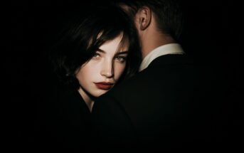 A woman with pale skin and dark hair gazes directly at the camera while embracing a man in a suit, whose back is turned. The background is dark, highlighting her face with dramatic lighting.