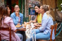 Four women sit around a table at an outdoor café, enjoying drinks and pastries while chatting and laughing together on a sunny day. A bicycle is parked nearby.