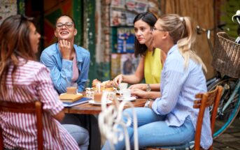 Four women sit around a table at an outdoor café, enjoying drinks and pastries while chatting and laughing together on a sunny day. A bicycle is parked nearby.