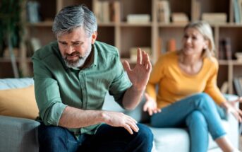 A middle-aged man sits on a couch with a distressed expression, raising his hand as if to stop the conversation, while a woman behind him gestures and appears to be speaking emotionally.