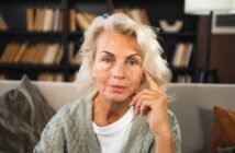 A mature woman with light blonde hair and blue eyes sits on a sofa, resting her hand on her face. She wears a gray sweater and white top, with a bookshelf full of books in the background.