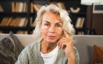 A mature woman with light blonde hair and blue eyes sits on a sofa, resting her hand on her face. She wears a gray sweater and white top, with a bookshelf full of books in the background.