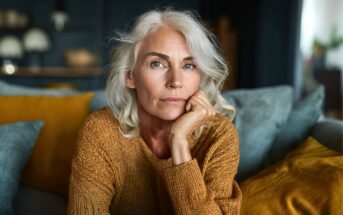 An older woman with gray hair and a thoughtful expression sits on a couch, resting her chin on her hand. She wears a textured brown sweater, and colorful cushions are in the background.