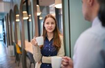 A young woman holding a white mug stands in a modern office hallway, talking to a person off to the right who is also holding a mug. She looks engaged in the conversation.