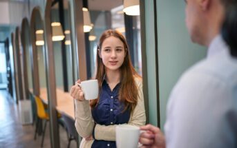 A young woman holding a white mug stands in a modern office hallway, talking to a person off to the right who is also holding a mug. She looks engaged in the conversation.