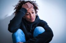 A person in a black hoodie and blue jeans sits against a white wall, looking down with a hand on their forehead, appearing sad or stressed.