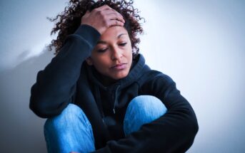 A person in a black hoodie and blue jeans sits against a white wall, looking down with a hand on their forehead, appearing sad or stressed.