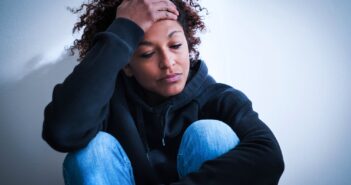 A person in a black hoodie and blue jeans sits against a white wall, looking down with a hand on their forehead, appearing sad or stressed.