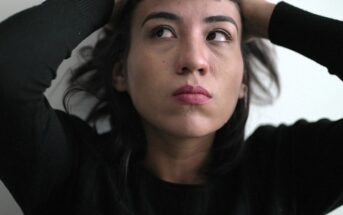 A person with dark hair and a serious expression looks upward, holding their hands on their head. They are wearing a black top, and the background is plain and light-colored.