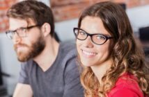 A woman with long wavy hair and glasses smiles at the camera, while a man with a beard and glasses sits beside her, slightly out of focus. They appear to be indoors in a casual setting.