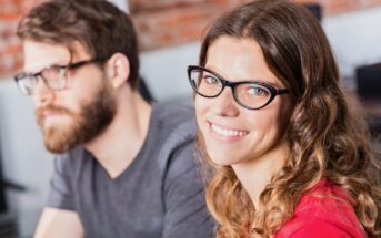 A woman with long wavy hair and glasses smiles at the camera, while a man with a beard and glasses sits beside her, slightly out of focus. They appear to be indoors in a casual setting.
