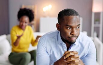 A man sits in the foreground looking thoughtful and concerned, with his hands clasped, while a woman in the background gestures emotionally, suggesting a serious conversation or disagreement between them.