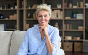 A smiling older woman with curly blonde hair sits on a light-colored sofa, resting her hand on her chin. She wears a light blue shirt. Bookshelves filled with books and decor are visible in the background.