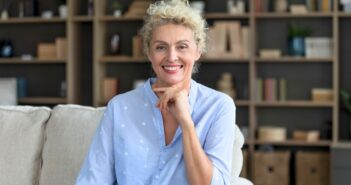 A smiling older woman with curly blonde hair sits on a light-colored sofa, resting her hand on her chin. She wears a light blue shirt. Bookshelves filled with books and decor are visible in the background.