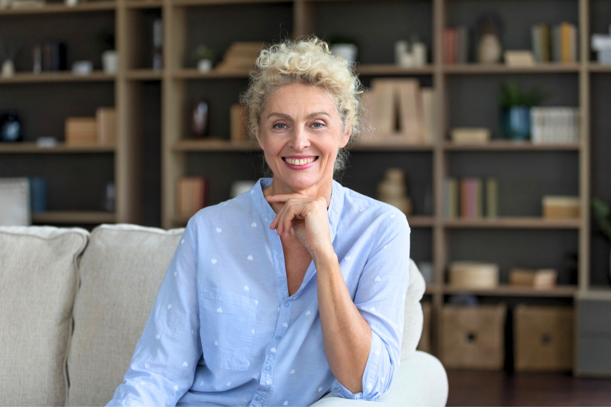 A smiling older woman with curly blonde hair sits on a light-colored sofa, resting her hand on her chin. She wears a light blue shirt. Bookshelves filled with books and decor are visible in the background.