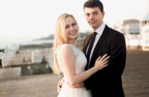 A smiling couple embraces on a wooden boardwalk. The woman wears a white sleeveless dress, and the man is in a black suit and tie. The background is bright and outdoors, with blurred railings and structures.