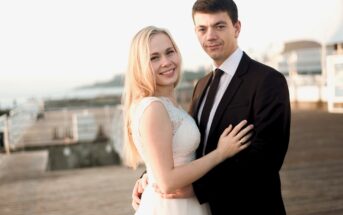 A smiling couple embraces on a wooden boardwalk. The woman wears a white sleeveless dress, and the man is in a black suit and tie. The background is bright and outdoors, with blurred railings and structures.