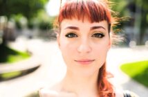 A young woman with red hair, winged eyeliner, and a nose ring looks directly at the camera outdoors on a sunny day, with a blurred background of green trees and a curved path.