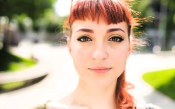 A young woman with red hair, winged eyeliner, and a nose ring looks directly at the camera outdoors on a sunny day, with a blurred background of green trees and a curved path.