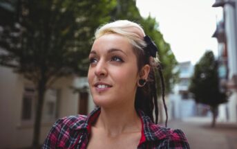 A person with light and dark hair styled in dreadlocks, wearing a plaid shirt and earrings, stands outdoors on a tree-lined street, looking slightly upward with a content expression.