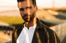 A man with dark hair and a beard stands outdoors, wearing a white shirt and a dark jacket. Sunlight casts a shadow across his face. The background is blurred, showing an outdoor walkway and distant structures.