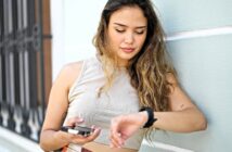 A young woman with long hair leans against a light-colored wall, holding a smartphone in one hand and looking down at her smartwatch on her wrist. She appears relaxed and focused.