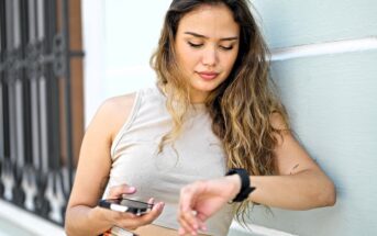 A young woman with long hair leans against a light-colored wall, holding a smartphone in one hand and looking down at her smartwatch on her wrist. She appears relaxed and focused.