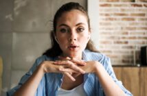 A young woman with light eyes and long brown hair, wearing a blue striped shirt, sits indoors with her hands clasped under her chin, looking attentively forward. A brick wall and tiled wall are in the background.