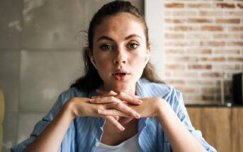 A young woman with light eyes and long brown hair, wearing a blue striped shirt, sits indoors with her hands clasped under her chin, looking attentively forward. A brick wall and tiled wall are in the background.