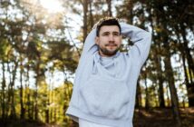 A man wearing a light gray hoodie stretches his arms over his head while standing outdoors in a sunny forest with trees in the background.