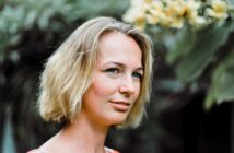 A woman with short blonde hair smiles softly outdoors, standing in front of green foliage and yellow flowers. The background is blurred, drawing attention to her face.