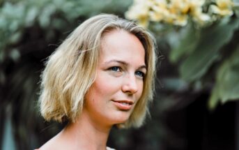 A woman with short blonde hair smiles softly outdoors, standing in front of green foliage and yellow flowers. The background is blurred, drawing attention to her face.