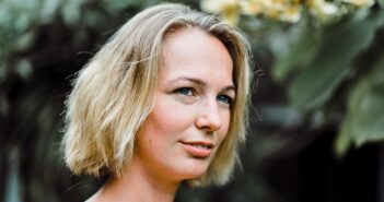 A woman with short blonde hair smiles softly outdoors, standing in front of green foliage and yellow flowers. The background is blurred, drawing attention to her face.