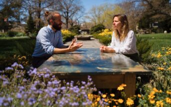 Two people sit across from each other at an outdoor table surrounded by blooming flowers and greenery, engaged in conversation on a sunny day with trees and a building in the background.
