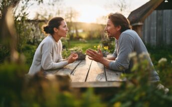 A man and woman sit across from each other at a wooden outdoor table, engaged in a serious conversation, surrounded by greenery, with warm sunlight in the background.