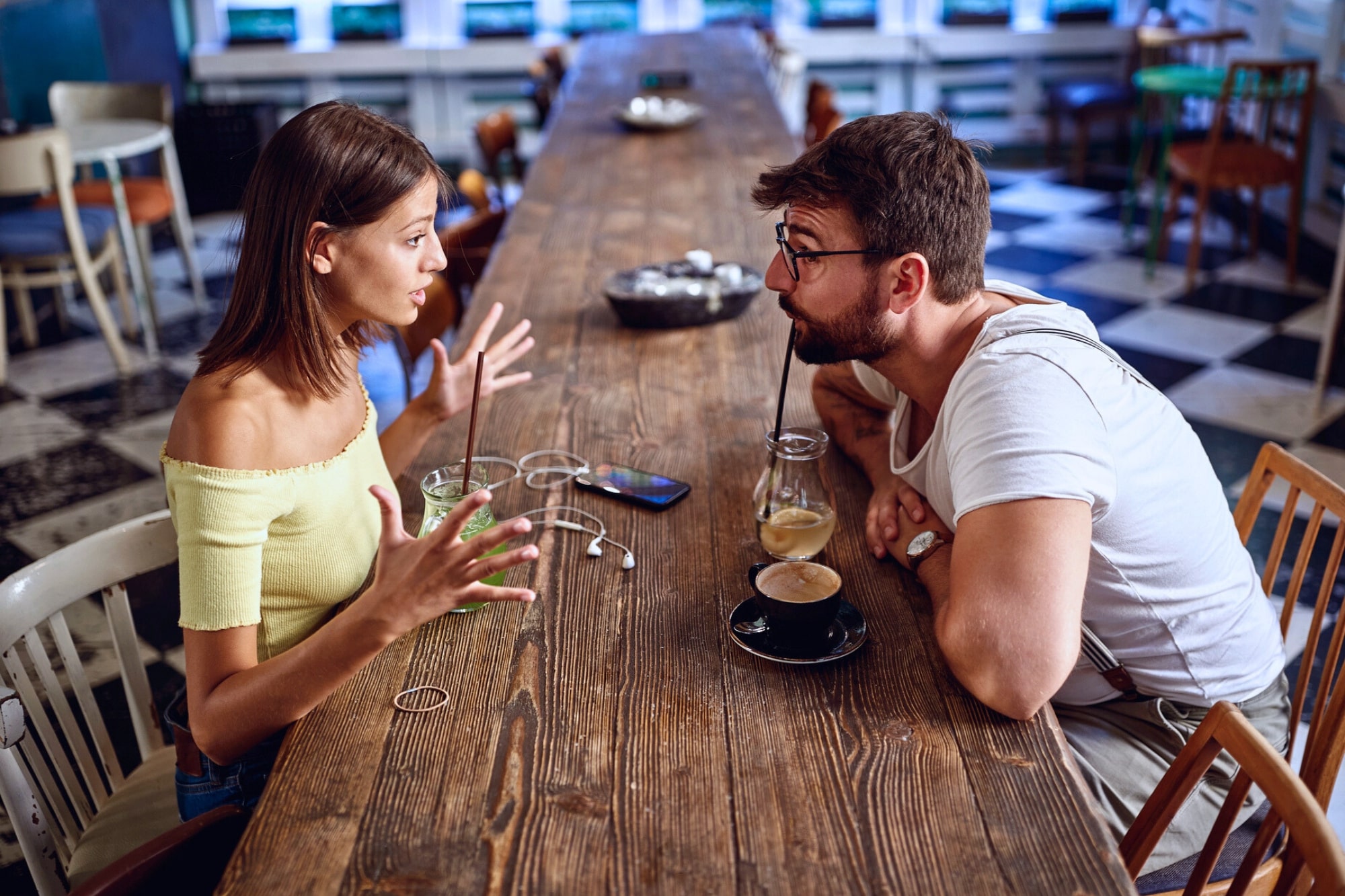 A woman and a man sit across from each other at a long wooden table in a cafe, engaged in an animated conversation, with drinks, a phone, and earphones on the table between them.