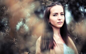 A young woman with long brown hair stands outdoors in front of blurry foliage, looking calmly at the camera. She wears a light-colored coat over a striped shirt. The image has a soft, natural, and slightly muted tone.