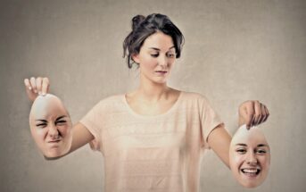 A woman looks down, holding two removable face masks in each hand—one with a scrunched, angry expression, the other with a big, happy smile. She stands against a plain, neutral background.